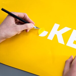 Puede incluir: Una mano utiliza un cuchillo de manualidades para despegar una pegatina amarilla que revela la palabra "OKE" en letras blancas sobre un fondo amarillo.