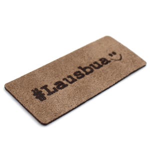 Puede incluir: Un parche de cuero marrón con el texto "#lausbua" y una cara sonriente.