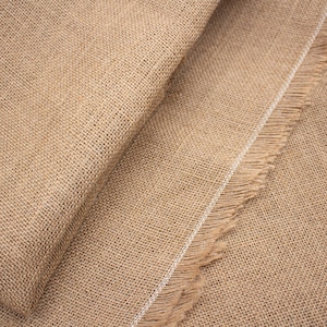 Può includere: Primo piano di un pezzo di tessuto di iuta naturale piegato. Il materiale ruvido e tessuto è di colore marrone chiaro, con una trama visibile e un bordo sfilacciato. Una striscia bianca corre lungo il bordo del tessuto.
