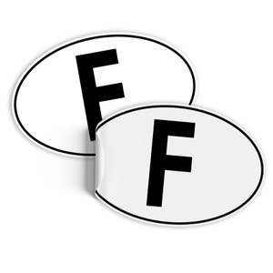 Può includere: Due adesivi ovali bianchi con bordo nero e una grande "F" nera al centro. Un adesivo è parzialmente staccato, rivelando l'adesivo. Gli adesivi sono per l'identificazione internazionale dei veicoli.