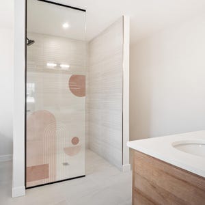 Op de afbeelding: Een moderne badkamerdouche met een glazen deur met een geometrisch ontwerp in roze en witte tinten. De doucheruimte heeft lichtgekleurde tegelwanden en een douchekop. Een houten wastafel met een wit aanrechtblad is zichtbaar.