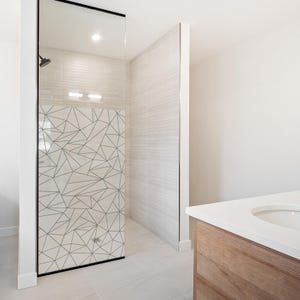 Könnte beinhalten: Ein modernes Badezimmer mit einer Glastür für die Dusche mit einem geometrischen Muster. Der Duschbereich hat hellfarbene Fliesen und einen schwarzen Duschkopf. Ein Holzwaschtisch mit weißer Arbeitsplatte ist sichtbar.