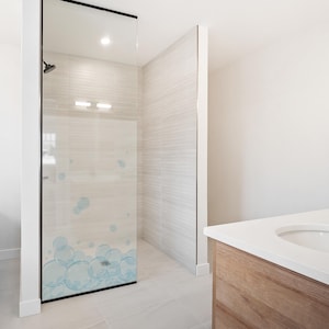 Könnte beinhalten: Modernes Badezimmer mit einer Glas-Duschkabine. Die Glastür hat ein blaues Blasendesign. Der Duschbereich hat hellfarbene Fliesenwände und einen Duschkopf. Eine weiße Arbeitsplatte mit Waschbecken ist sichtbar.