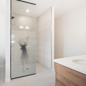 Könnte beinhalten: Eine Duschkabine mit einer Glaswand, die eine mattierte Hirschsilhouette zeigt. Die Dusche hat einen schwarzen Metallrahmen, helle horizontale Wandfliesen und einen an der Wand montierten Duschkopf im Inneren des gefliesten Bereichs.