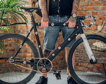 Könnte beinhalten: Ein schwarzes Fahrrad mit goldener Kette und schwarzen Reifen. Der Fahrradrahmen hat silberne Zierelemente. Der Lenker hat ein schwarz-weißes Muster. Das Fahrrad wird vor einer Backsteinmauer gezeigt.