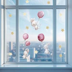 Könnte beinhalten: Fensteraufkleber mit weißen Hasen, die rosafarbene Ballons mit weißen Punkten halten. Die Hasen fliegen vor dem Hintergrund einer Stadtkulisse. Goldene Sterne sind über das Design verteilt.