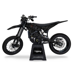 Könnte beinhalten: Schwarz-graues Dirtbike mit einem schwarz-weißen Tarnmuster. Das Bike steht auf einem schwarzen Ständer mit dem Wort "Racing" darauf.