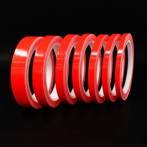 Könnte beinhalten: Ein Satz mit sechs roten doppelseitigen Klebebandrollen. Die Rollen sind übereinander gestapelt, wobei das Klebeband nach außen zeigt.