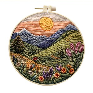 May include: Embroidered hoop art depicting a sunset over a mountain range with a green valley and wildflowers. The hoop is made of wood and has a natural finish.