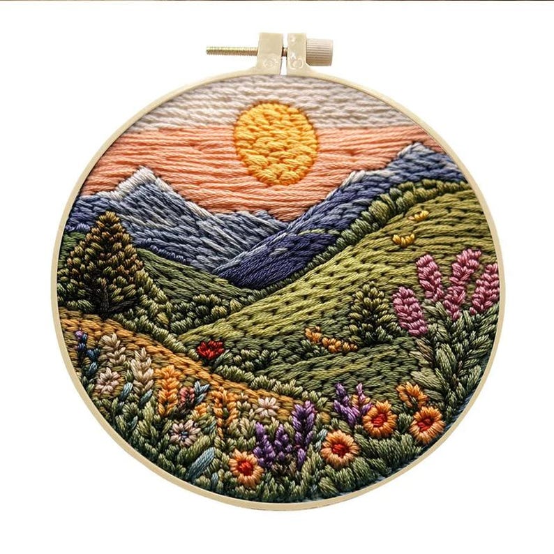 May include: Embroidered hoop art depicting a sunset over a mountain range with a green valley and wildflowers. The hoop is made of wood and has a natural finish.