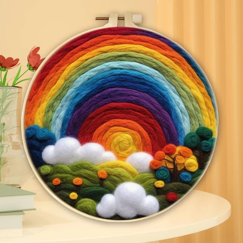 May include: A colourful rainbow made of yarn is displayed in a wooden embroidery hoop. The rainbow is surrounded by white clouds and green hills with small yellow flowers.