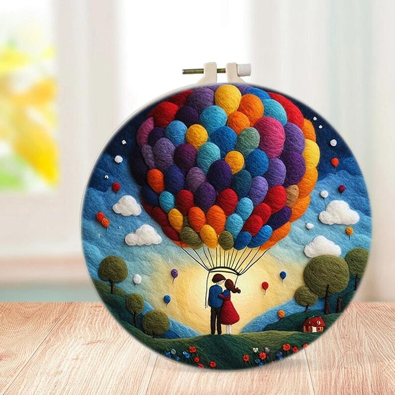 May include: A colourful felt embroidery hoop art piece depicting a couple standing under a hot air balloon made of colourful felt balls. The background is a blue sky with white clouds and green hills.