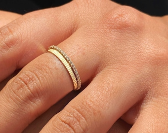 Può includere: Due anelli d'oro, uno con un design strutturato e scanalato e l'altro con piccole pietre preziose trasparenti. Gli anelli sono indossati su un dito, mostrando i loro dettagli delicati e lo stile elegante. Gli anelli sono un gioiello classico.