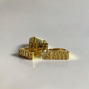 Op de afbeelding: Drie gouden ringen met de woorden "SHAKE", "GRUNGE" en "SLAYYYY" erop gegraveerd.