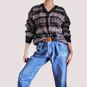 May include: A brown, gray, and white patterned sweater with a v-neck and long sleeves. The sweater is worn over a pair of blue jeans with a brown leather belt.