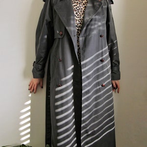 May include: A long, dark gray trench coat with a double-breasted closure and a striped pattern. The coat has a classic design with a collar and pockets.