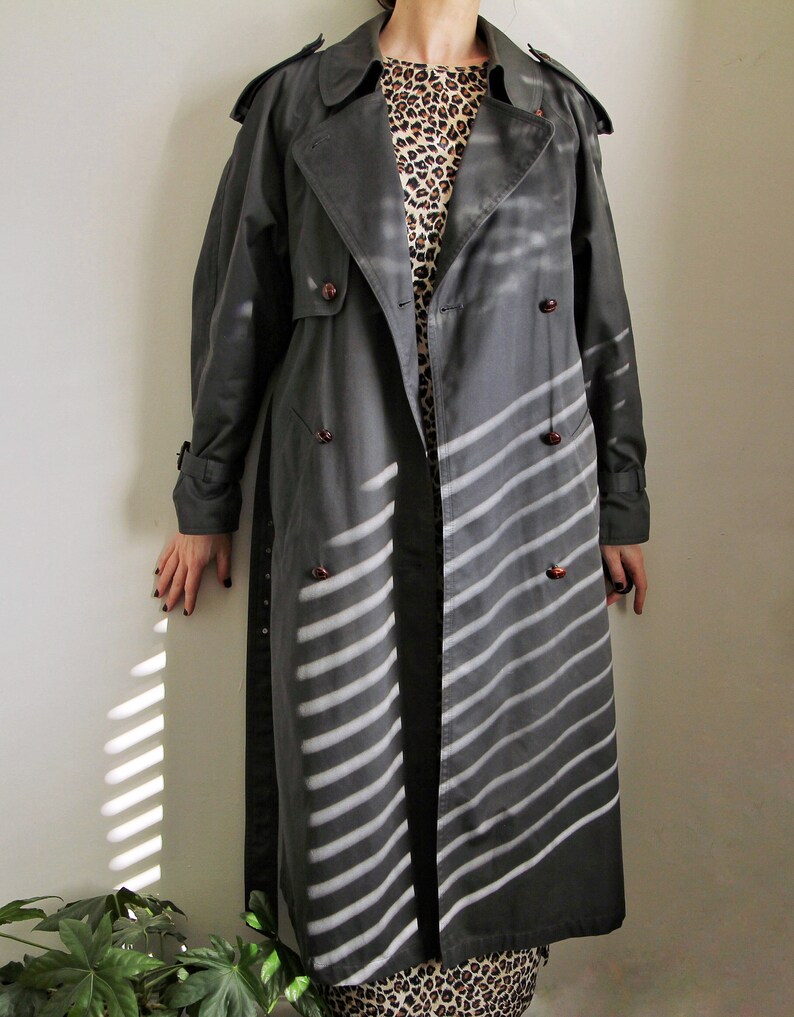 May include: A long, dark gray trench coat with a double-breasted closure and a striped pattern. The coat has a classic design with a collar and pockets.