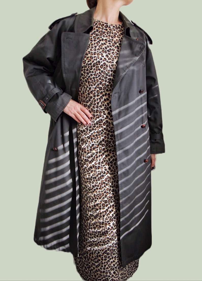 May include: A long, gray trench coat with white stripes. The coat has a double-breasted closure and a belt. The coat is worn over a leopard print dress.