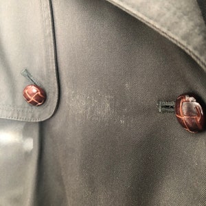 May include: Close-up of a dark gray fabric with two brown buttons. The buttons have a woven design and are sewn onto the fabric.