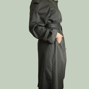 May include: A long, dark green trench coat with a belt and a button closure. The coat has a classic design with a collar and pockets.