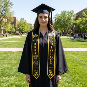 May include: A black graduation gown and cap with a black tassel, worn with two black graduation stoles featuring gold embroidery. The stoles display the text 'CLASS OF 2026', 'CUSTOM NAME', 'RADIOLOGY', and 'RT', along with a graduation cap icon and a radiation symbol.