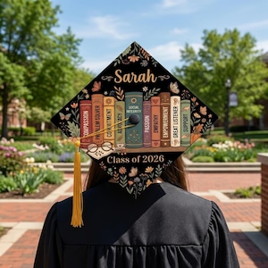 May include: A black graduation cap topper featuring a row of colorful books with titles like Compassion, Commitment, and Social Integrity. The name Sarah and Class of 2026 are printed in white. A yellow tassel hangs from the center button.