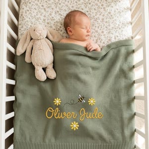 May include: A sage green knitted baby blanket featuring the embroidered name Oliver Jude in yellow script, surrounded by three yellow daisy flowers and a small bee with a dotted flight path, accompanied by a beige plush rabbit toy.