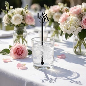 May include: A clear glass tumbler filled with ice cubes and a black acrylic drink stirrer featuring the letters I & A at the top. The stirrer is positioned vertically inside the glass, which sits on a white tablecloth near floral arrangements.