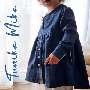 May include: A blue denim tunic with long sleeves and a button-up front. The tunic has a gathered skirt and a white trim on the sleeves. The text "Ebook Tunika Mika Gr. 86 - 140" is written in white on the left side of the image.