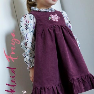 May include: A burgundy corduroy dress with a floral embroidery detail on the bodice. The dress has a ruffled hem and is worn over a white floral print blouse. The dress is labeled "Trägerkleid Freige Gr. 86 - 140".
