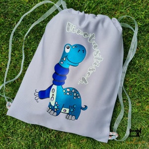 May include: Gray drawstring bag with a blue dinosaur wearing a blue scarf. The bag has the text "Dino tastisch" on it.