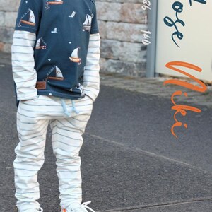 May include: A young boy wearing a blue and white striped long-sleeve shirt with a sailboat print and white striped pants. The text "Hose Niki Gr. 86 - 140" is visible in the image.