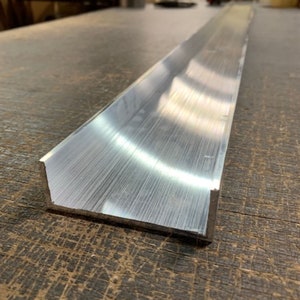 Puede incluir: Un canal en forma de U de aluminio plateado, con acabado cepillado, se encuentra sobre una superficie de madera.