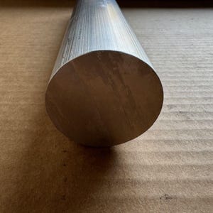以下が含まれることがあります： 円筒形の金属棒で、端は円形です。棒は銀色で、アルミニウムまたは同様の合金でできているようです。棒の端は滑らかで、金属の木目が見えます。棒は茶色の表面に置かれています。
