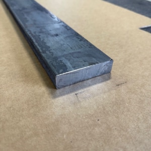 May include: A long, rectangular piece of flat steel bar stock. The bar is gray and has a rough, unfinished surface.