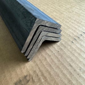 May include: Close-up of several steel angle bars, each with a 90-degree bend. The bars are stacked, revealing their cut edges. The metal has a dark, industrial finish, and the bars are resting on a brown surface.