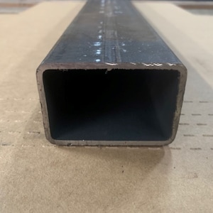 May include: A close-up of a black metal square tube. The tube is hollow and has a smooth, slightly textured surface.