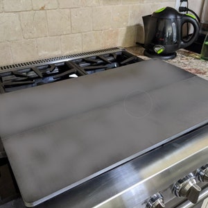 May include: A black metal stovetop cover sits on a stainless steel stovetop. The cover is flat and has a slight curve to it. The stovetop has four knobs on the right side.
