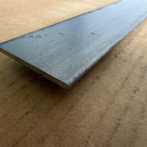 May include: A flat, rectangular piece of unfinished steel. The metal has a matte gray finish and is lying on a brown surface. The steel bar is likely used in construction or metalworking projects.