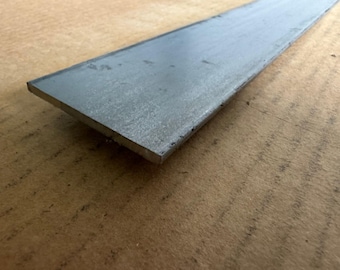 May include: A flat, rectangular piece of unfinished steel. The metal has a matte gray finish and is lying on a brown surface. The steel bar is likely used in construction or metalworking projects.