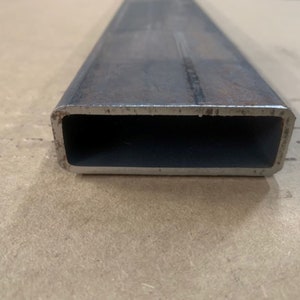 May include: Close-up of a rectangular steel tube with a slightly rusted surface. The tube is hollow and has a smooth, polished interior.