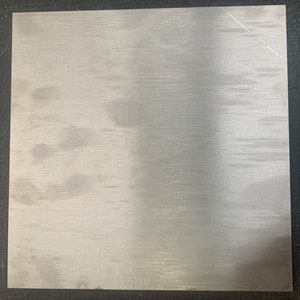 Puede incluir: Una placa cuadrada de metal cepillado con acabado plateado. La superficie tiene un patrón de grano lineal que corre horizontalmente. La placa está sobre un fondo oscuro.