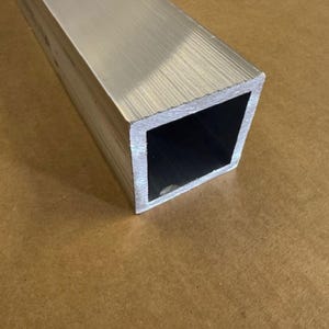 May include: A close-up of a silver square aluminum tube. The tube has a brushed finish and is lying on a brown surface.