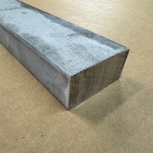 May include: A rectangular bar of metal with a rough, gray surface. The bar is slightly weathered and has a subtle sheen.