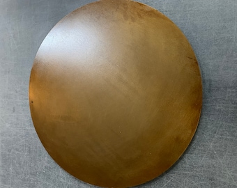 May include: A large, circular, bronze-colored metal plate. The plate has a smooth surface with subtle variations in tone, suggesting a brushed or aged finish. The overall appearance is simple and industrial.