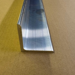 May include: A close-up of a brushed aluminum L-shaped metal piece. The metal has a reflective surface and is set against a tan background. The piece has a rounded edge.