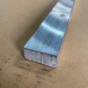 May include: A rectangular, silver-colored metal bar with visible machining marks. The bar has a rough, textured end and appears to be made of steel or a similar alloy. The surface shows some wear and tear.
