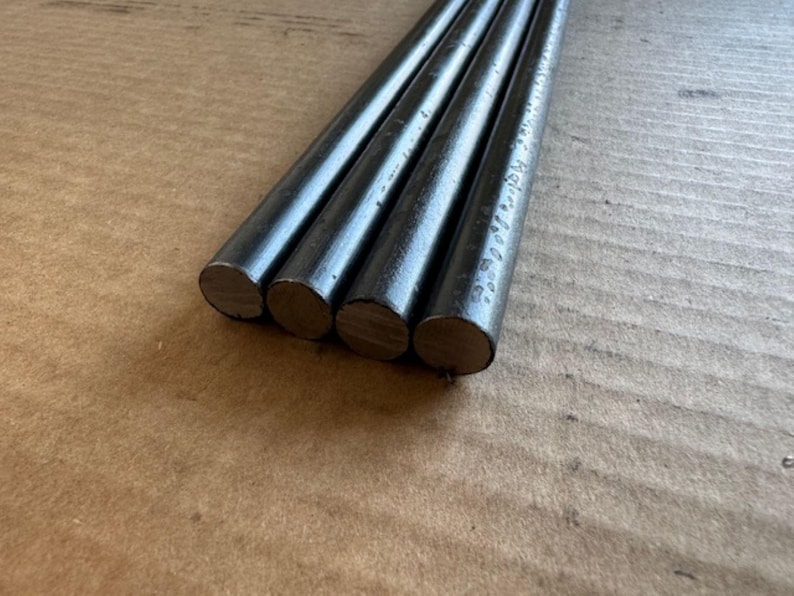 May include: Four dark grey metal rods with rounded ends are arranged diagonally. The rods have a slightly textured surface and appear to be made of steel. The rods are lying on a brown surface.