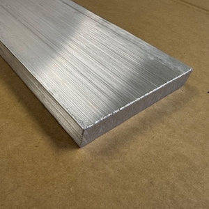 May include: A rectangular, brushed aluminum bar with a silver finish. The metal bar has a textured surface with visible linear graining. The bar is displayed on a brown surface.
