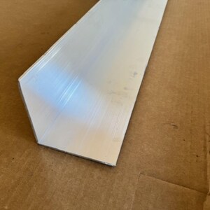 May include: A silver aluminum angle, or corner brace, with a brushed finish. The angle is made of metal and has a 90-degree angle.